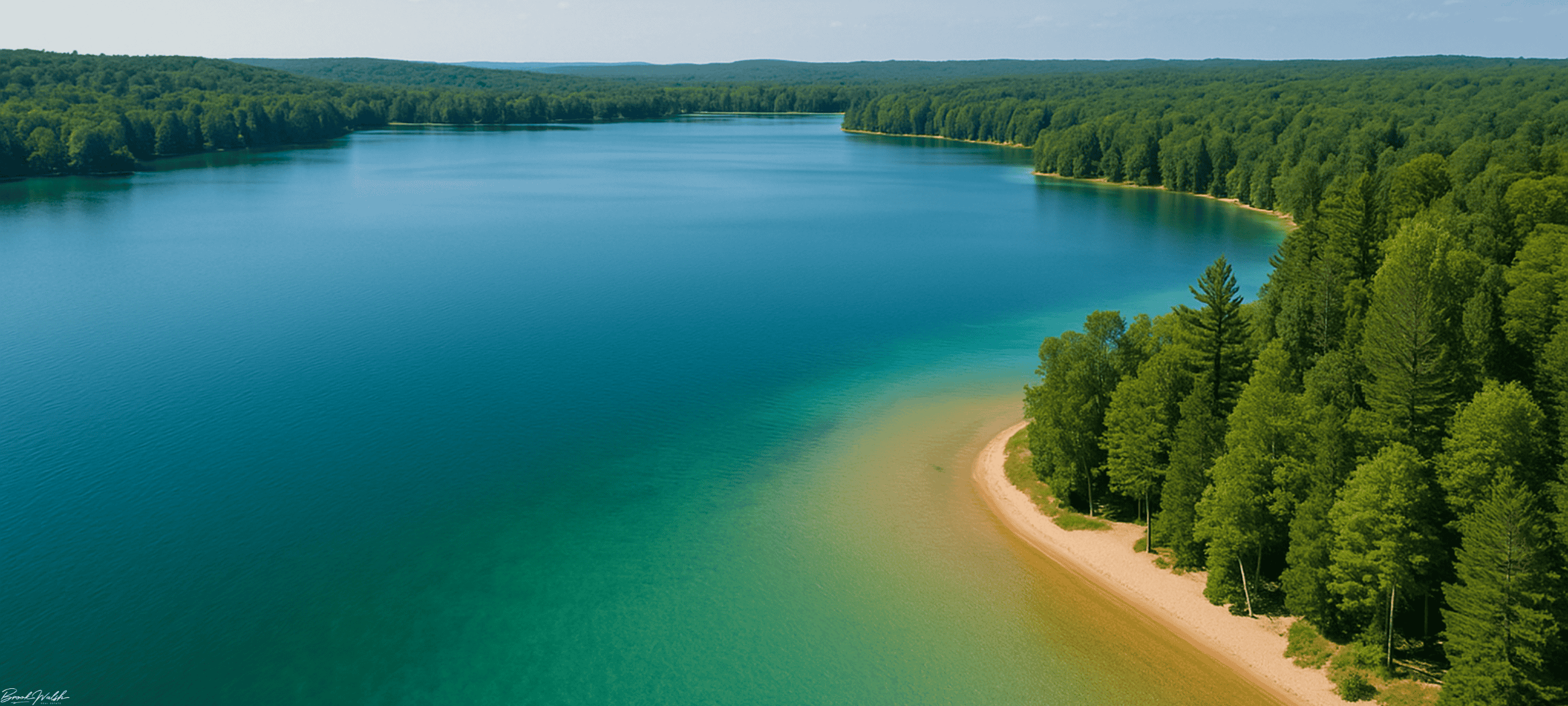 Aerial view of Deer Lake’s clear waters, sandy shoreline, and surrounding northern Michigan forest in summer.